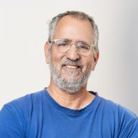 גבר עם זקן אפור וחולצה כחולה מחייך למצלמה.