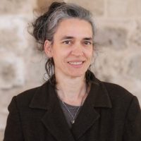 אישה עם שיער אפור במעיל כהה, מחייכת מול קיר אבן.