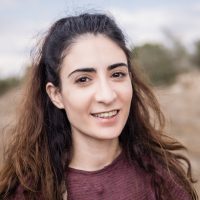 אישה צעירה עם שיער ארוך וגולש מחייכת למצלמה, לובשת חולצה מפוספסת, עם רקע טבעי מטושטש.