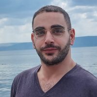 גבר עם משקפיים וזקן עומד ליד אגם, לובש חולצה כהה, עם הרים ברקע.