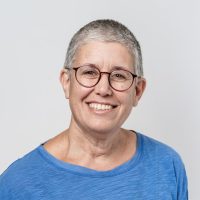 אישה מחייכת עם שיער אפור קצר ומשקפיים, לובשת חולצה כחולה.